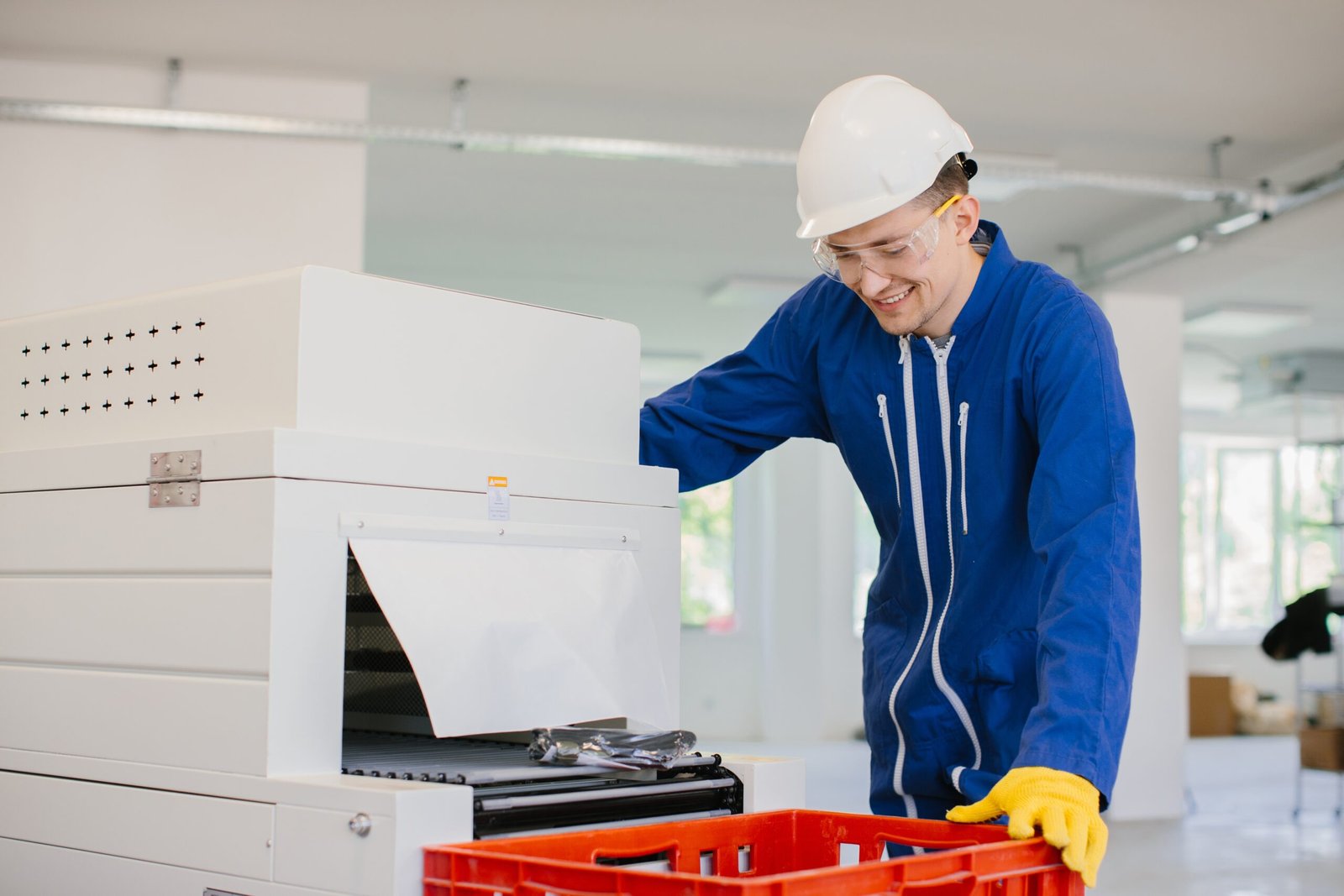 Factory worker wearing safety glasses and hardhat operating industrial machinery and holding a red crate